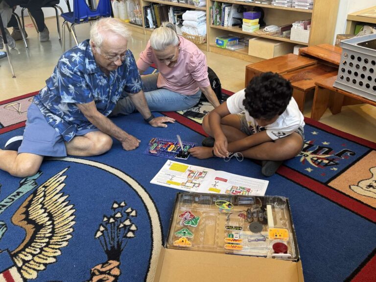 Grandparents sitting on the classroom rug, working on a Montessori lesson with a student