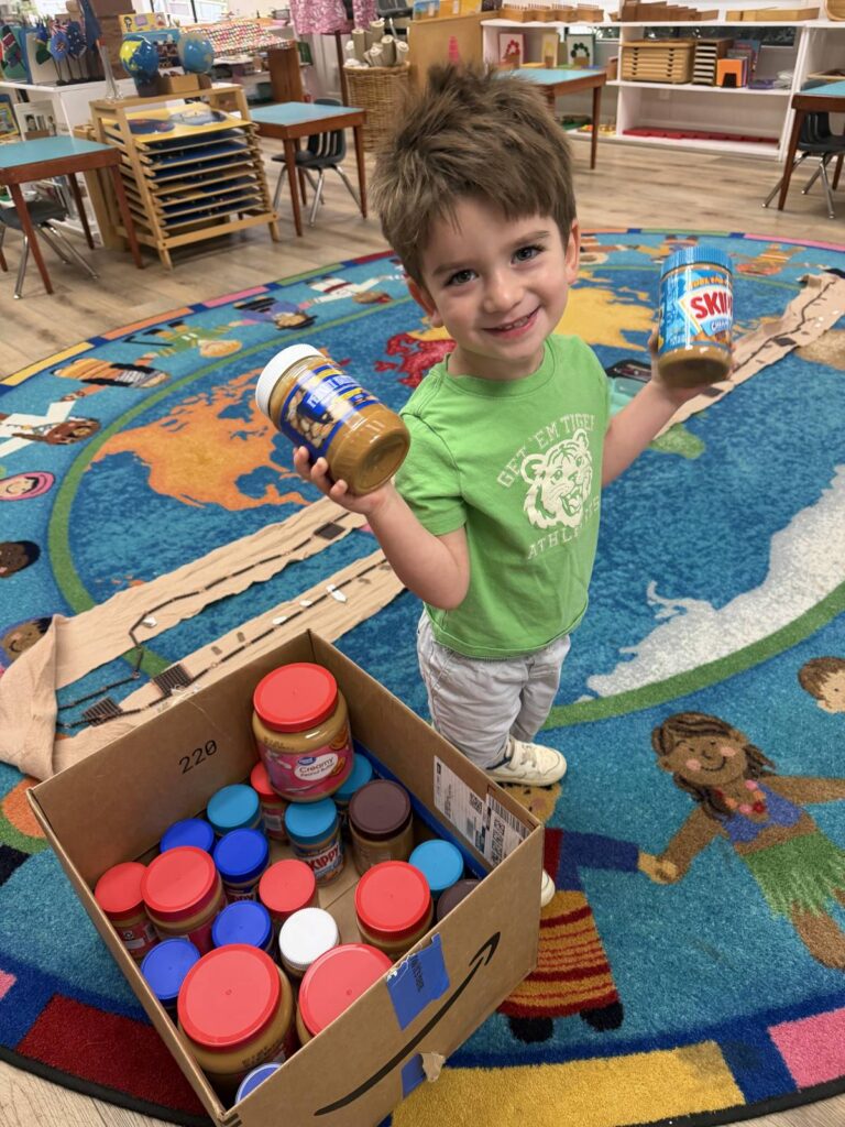 Preschool student holding jars of peanut butter beside a box of donations for Feeding San Diego